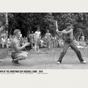 May include: A black and white historical photograph titled PHOTOGRAPH OF THE CHRISTMAS DAY BASEBALL GAME 1943. It depicts a baseball game in progress with a batter holding a wooden bat, a catcher in a crouched position, and spectators.
