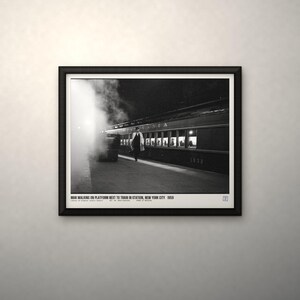 May include: A black and white framed photograph titled MAN WALKING ON PLATFORM NEXT TO TRAIN IN STATION, NEW YORK CITY 1959. The image depicts a person in a coat walking on a train platform beside a steam-filled passenger train car.
