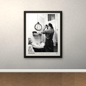 May include: A black and white framed photograph titled DOLLAC STEEL MILL. The image depicts a person standing at a bathroom sink grooming their hair while a child watches. A round mirror hangs above the sink, and a window with a curtain is visible.