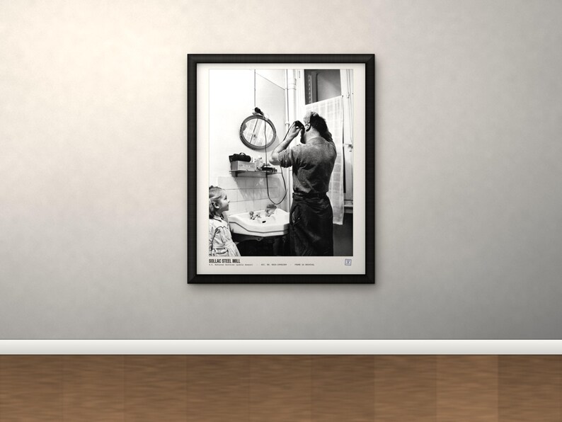 May include: A black and white framed photograph titled DOLLAC STEEL MILL. The image depicts a person standing at a bathroom sink grooming their hair while a child watches. A round mirror hangs above the sink, and a window with a curtain is visible.