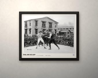 May include: A framed black and white photograph titled BOXING, CAMP CUSTER, MICH. The image depicts two boxers in a ring surrounded by a crowd of soldiers in front of wooden barracks. One boxer wears white shorts, the other wears dark trunks.