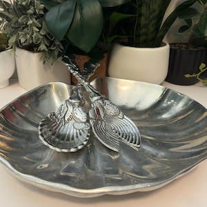Könnte beinhalten: Eine silberfarbene Servierschale in Form einer großen Jakobsmuschel, begleitet von einem passenden Salatbesteck. Die Griffe des Bestecks sind mit detaillierten Muschelreliefs verziert, während die Schale eine glatte, reflektierende Oberfläche aufweist.
