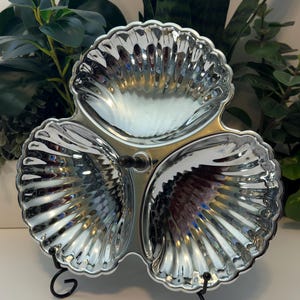 May include: A silver-toned metal serving dish featuring three interconnected shell-shaped bowls arranged in a clover pattern. The dish has a reflective, polished surface with scalloped edges and a small central handle, supported by a black wire stand.
