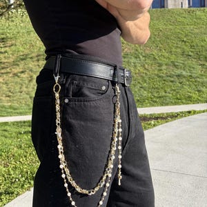 Puede incluir: Una cadena de cartera de tono dorado de múltiples hilos sujeta a un cinturón de cuero negro. El accesorio presenta una combinación de eslabones metálicos gruesos, hilos de cuentas delicados con pequeñas perlas blancas y pequeños dijes.