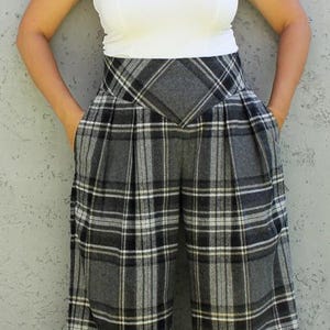 以下が含まれることがあります： グレー、黒、白のチェック柄のワイドパンツ。ハイウエストのパンツにはプリーツが入っています。白いタンクトップと明るい色の靴を履いています。
