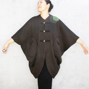 以下が含まれることがあります： Vネックのダークブラウンのゆったりとしたポンチョ。フロント中央には、黒い布製の水平な留め具が3つあり、それぞれに丸いゴールドトーンのボタンが付いています。前面の両サイドには、大きな楕円形のパッチポケットが2つ付いています。