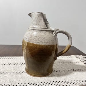 May include: A ceramic pitcher featuring a two-tone glaze design. The upper portion is a speckled off-white, while the lower section is a solid, glossy brown. It includes a curved handle and a distinct pouring spout at the top rim.