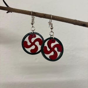 Puede incluir: Un par de pendientes colgantes con un marco circular de color verde oscuro que encierra un diseño de cruz curva de cuatro brazos en color rojo. Los pendientes están sujetos a ganchos de metal plateado y cuelgan de una rama marrón.