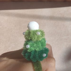 May include: A ring featuring a small, tiered Christmas tree crafted from green pipe cleaners. The tree is decorated with tiny white beads and topped with a white pom-pom. The band of the ring is made from a single green pipe cleaner.