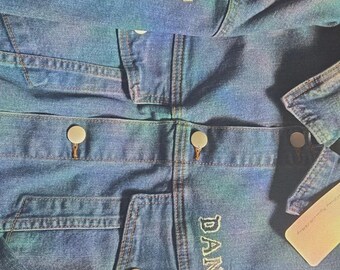 May include: A blue denim jacket with an iridescent finish, featuring the text 1987 embroidered on the upper sleeve and the name DANIEL embroidered above the chest pocket. The jacket includes metal buttons and a tan paper tag attached.