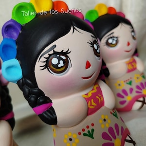 Puede incluir: Dos figuras de cerámica que representan un personaje con cabello negro trenzado, adornado con una diadema de colores del arcoíris. Las figuras tienen ojos grandes de color marrón, una nariz roja pequeña y un vestido beige con patrones florales amarillos y morados. El texto Taller de los Sueños es visible.