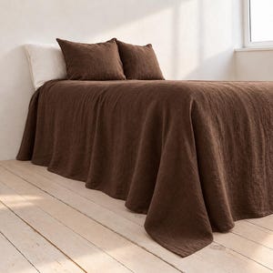 May include: A dark brown bedspread with a textured finish covers a bed, accompanied by two matching dark brown square pillow shams and one plain white pillow, all arranged neatly on a bed frame in a bright, sunlit room setting.