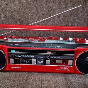 Könnte beinhalten: Ein leuchtend roter tragbarer Sanyo Stereo-Kassettenrekorder mit zwei Kassettendecks mit Auto-Reverse-Funktion, zwei großen schwarzen Lautsprechern mit silbernen Rändern, einer langen silbernen Teleskopantenne und einem roten Tragegriff.