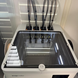 May include: A white Snapmaker 3D printer featuring a clear top enclosure with a black frame. The unit includes four vertical filament tubes extending upward, a central circular Snapmaker logo on the lid, and the brand name printed on the front panel.