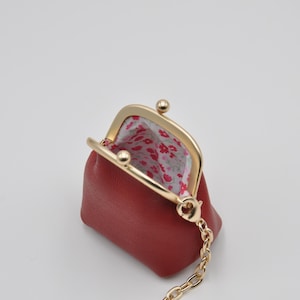 Op de afbeelding: Een klein rood leren muntportemonnee met een gouden metalen frame en een ketting eraan. De portemonnee is open en de binnenkant is gevoerd met een witte stof met een roze bloemenpatroon.