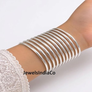 May include: A set of ten smooth, polished silver-toned bangles worn together on the wrist. The bracelets are thin, circular, and have a reflective metallic finish. The text JewelsIndiaCo is visible in the lower portion of the image.