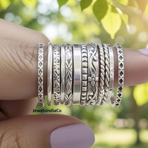 Puede incluir: Un conjunto de diez anillos de tono plateado con diversas texturas y patrones, incluyendo bandas lisas, diseños de cuerda trenzada, motivos florales y formas geométricas, alineados en un dedo con el texto JewelsIndiaCo.