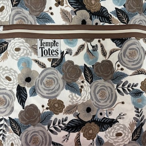 Peut inclure: Une pochette zippée en tissu blanc avec une fermeture éclair marron et une bordure marron. Le tissu présente un motif floral avec des fleurs grises, brunes et bleues. La pochette porte une étiquette indiquant "Temple Totes".