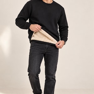 May include: A black long-sleeve sweatshirt with a beige faux-fur lining, paired with dark gray denim jeans and white sneakers. The sweatshirt features a crew neckline, ribbed cuffs, and a hem that reveals the thick, textured interior lining.