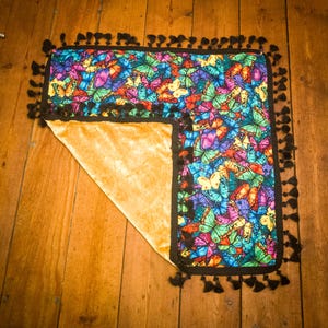 May include: A square textile piece featuring a vibrant, multi-coloured butterfly pattern on one side and a solid gold-toned velvet fabric on the other. The edges are finished with a black border and a series of small, dangling black tassels.