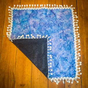 May include: A square fabric piece featuring a blue and purple galaxy-inspired print with white speckles. The edges are trimmed with a silver metallic decorative border adorned with dangling clear teardrop-shaped beads. One corner is folded over.