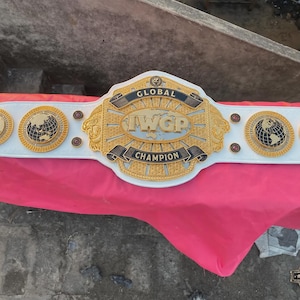 Könnte beinhalten: Ein weißer Wrestling-Meisterschaftsgürtel aus Leder mit einer großen, zentralen goldenen Platte, die den Text GLOBAL IWGP CHAMPION trägt. Der Gürtel enthält vier kleinere kreisförmige goldene Platten mit Weltkarten.