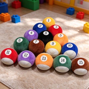 May include: A set of fifteen plush billiard balls arranged in a triangle formation. Each soft ball features a distinct color and a white circular patch with a black number from one to fifteen printed in the center, mimicking standard pool balls.