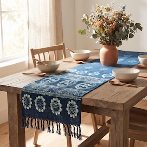 May include: A rectangular blue table runner featuring a white floral and geometric block-print pattern with a fringed edge. The runner is draped across a wooden table set with ceramic bowls, wooden spoons, and a vase of dried flowers.