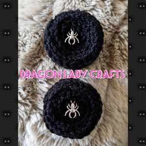 May include: Two black crocheted circles with a silver spider charm in the centre of each. The circles are on a fluffy white surface. The text "DragonLady Crafts" is in pink letters in the centre of the image.