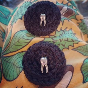 May include: Two black crocheted circles with a silver tooth charm in the center of each. The circles are on a yellow and green fabric background.