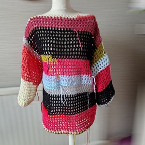 May include: A colorful striped crochet top with a loose fit and a wide neckline. The top is made with a variety of colors, including pink, black, yellow, blue, and gray. The top has a crocheted texture and is finished with fringe details.