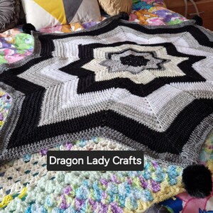May include: A black, white, and gray crocheted star-shaped blanket with a white center and black pom-poms on the corners. The blanket is on a bed with a colorful crocheted blanket underneath. The text "Dragon Lady Crafts" is visible in the lower center of the image.