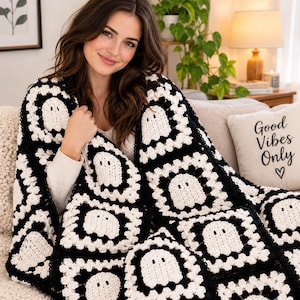 May include: A handmade crochet blanket featuring a repeating pattern of white ghost motifs on black and white granny squares. The blanket is constructed from individual square blocks joined together, creating a textured, repeating design.