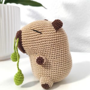 Könnte beinhalten: Ein beiges, gehäkeltes Capybara-Stofftier mit dunkelbraunen Ohren, Füßen und Schnauze. Die Figur hat ein mit schwarzem Faden gesticktes geschlossenes Auge und hält einen grünen Stängel mit zwei kleinen grünen Kugeln, die aus dem Mund hängen.