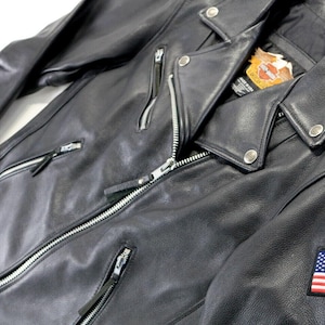 May include: A black leather motorcycle jacket featuring silver-toned metal zippers, snap button lapels, and a Harley-Davidson brand label on the inner collar. An embroidered American flag patch is stitched onto the left sleeve of the garment.