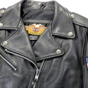 May include: A black leather motorcycle jacket featuring silver-tone metal zippers, snap buttons on the lapels, and shoulder epaulets. An American flag patch is on the sleeve, and the interior label reads Harley-Davidson An American Legend.