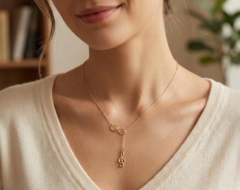 May include: A gold-colored necklace featuring an infinity symbol and a treble clef pendant. The necklace is worn on a person with a cream-colored v-neck sweater. The background is blurred, with a hint of a bookshelf and a plant.