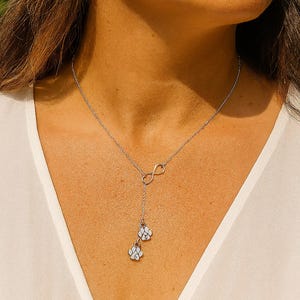 May include: A silver necklace with an infinity symbol pendant and a drop chain holding two white paw print charms. The necklace is worn over a white blouse.