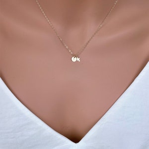 May include: A delicate gold-colored necklace with a small pendant featuring a round charm and a cross. The necklace is displayed against a white top, highlighting the jewelry's design. The chain is fine and elegant.