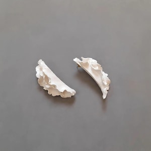 Könnte beinhalten: Ein Paar weiße Ohrstecker in einer geschwungenen, länglichen Form mit zart gewellten Rändern. Die Oberfläche der Ohrringe ist glatt und matt und zeigt ein geschichtetes, wellenartiges Design, das ein Gefühl von Bewegung erzeugt.