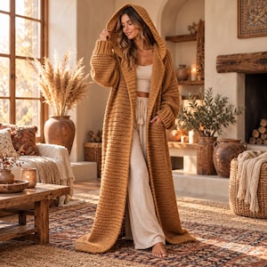 May include: A long, floor-length, mustard-yellow chunky knit cardigan featuring a hood, long sleeves with ribbed cuffs, and two front pockets. The garment is worn over a light beige tank top and matching wide-leg trousers with a drawstring waist.