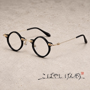 Puede incluir: Gafas redondas con monturas circulares negras, un puente de metal dorado y varillas finas doradas con puntas negras. Las gafas incluyen lentes transparentes y almohadillas nasales ajustables. El texto "こばやしげんぞう" es visible en la esquina inferior derecha.