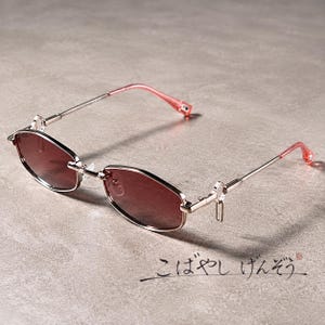 Puede incluir: Gafas de sol con montura metálica fina y lentes hexagonales en un tono marrón rojizo oscuro. Las varillas son metálicas con puntas de color rosa translúcido. El texto japonés "こばやし げんぞう" es visible en la parte inferior derecha.