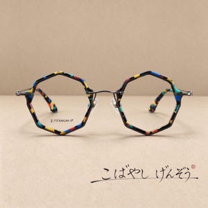 May include: Octagonal spectacles featuring a multicoloured patterned frame with blue, yellow, and red accents. The thin metal bridge connects the rims, and the lens is marked with the text β-TITANIUM-IP. The temple arms display a matching pattern.