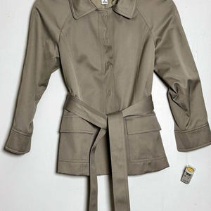 May include: A light brown, long-sleeved jacket featuring a pointed collar, a hidden button placket, and a matching fabric tie belt at the waist. The jacket includes two front flap pockets and a Talbots brand label at the inner neckline.