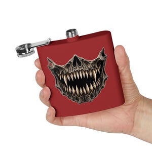 May include: A matte red metal flask featuring a graphic print of a menacing skull with sharp, pointed teeth. The flask has a silver-coloured screw-top lid attached to a hinged metal arm, designed for secure closure and easy access.