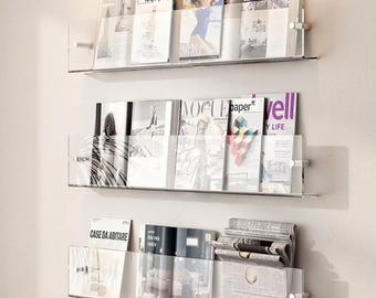 May include: Three clear acrylic wall-mounted floating shelves holding various magazines and newspapers. The shelves feature silver-toned metal hardware on the sides. The publications include titles like Wallpaper, Vogue, and Case Da Abitare.