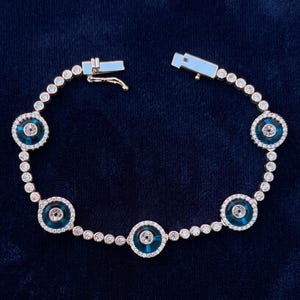 Könnte beinhalten: Ein goldfarbenes Tennisarmband mit fünf kreisförmigen blauen Schutzaugen-Anhängern, die mit kleinen klaren Steinen durchsetzt sind. Das Armband besteht aus einer Reihe runder, klarer Steine in Krappenfassung und hat einen sicheren Kastenschloss.