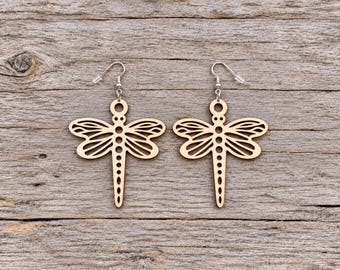 May include: A pair of light-colored wooden earrings shaped like dragonflies. Each earring features a laser-cut design with openwork wings and a segmented body, suspended from silver-toned metal fishhook wires with clear plastic earring backs.