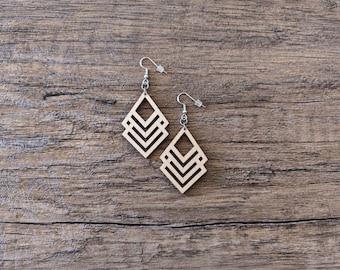 May include: A pair of light-colored wooden earrings featuring a geometric diamond shape composed of three nested chevron cutouts. Each earring is attached to a silver-toned metal fishhook ear wire with a small clear plastic backing for secure wear.